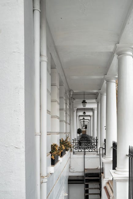 A covered outdoor corridor in a residential building with white walls and large white columns supporting the ceiling. Several black metal lantern-style light fixtures are mounted along the ceiling, and there are potted plants with dried flowers placed on the ledge to the left. The corridor extends into the distance, with a black metal fence and steps visible at the far end. The space is part of a home or building, potentially in a historic or upscale area such as Belgravia. This setting is characteristic of the architecture often involved in house removals and relocation services, where careful access considerations are needed for furniture transport and packing during a home relocation process, as managed by local removal companies like Removal Van Belgravia.