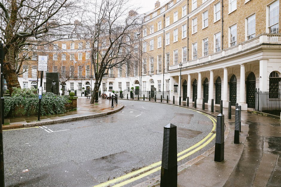 Street view of a narrow access residential area in Belgravia, featuring a curved, wet asphalt road bordered by black bollards and pavement. On the right, there is a large white building with tall, arched doorways protected by black iron gates, supported by white columns, and with multiple rows of tall windows. A tree without leaves stands near the sidewalk, with some green shrubbery and a park bench nearby. The scene is overcast with subdued natural light, indicating recent rain. The surroundings are clean and well-maintained, typical of an affluent Belgravia neighbourhood. The setting provides a realistic backdrop for house removals and furniture transport, highlighting the challenges of narrow access areas in property relocation in central London, as managed by Removal Van Belgravia, which specializes in efficient removals and packing services for homes in this area.