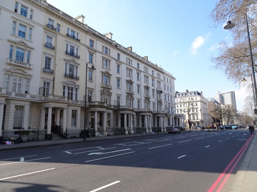 A wet, narrow street in Belgravia SW1X, featuring a curved road bordered by white, multi-story Victorian-style buildings with large windows and black wrought iron gates. The street includes a small tu